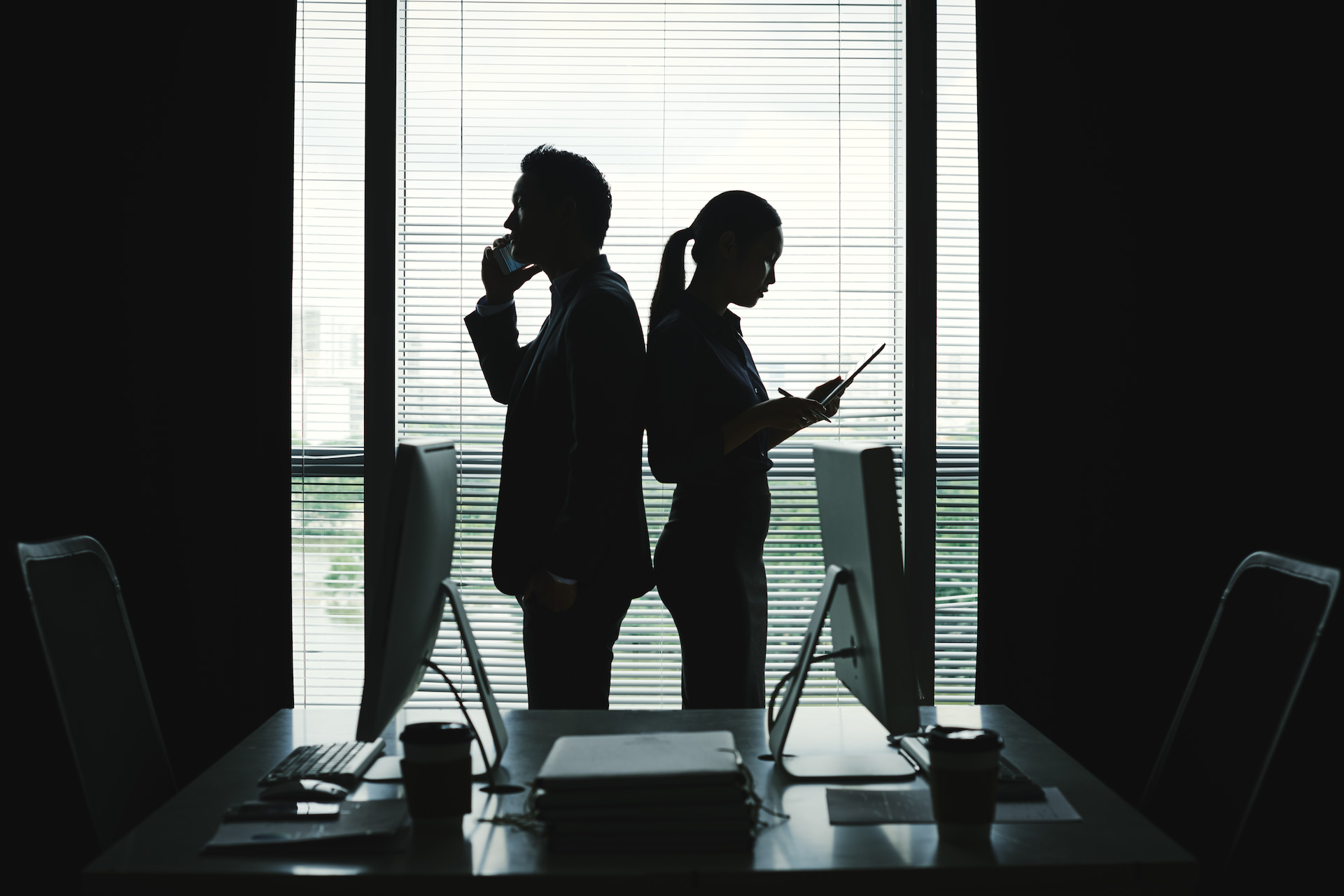 Two colleagues in silhouette at a bright office window, one on the phone