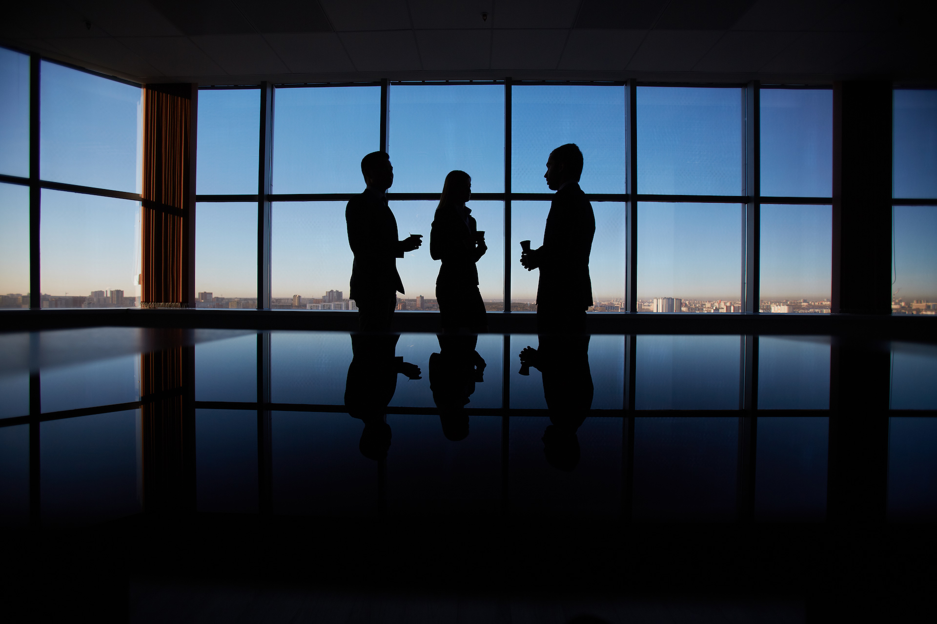 Three people in silhouette talking in front of a wall of windows at dusk