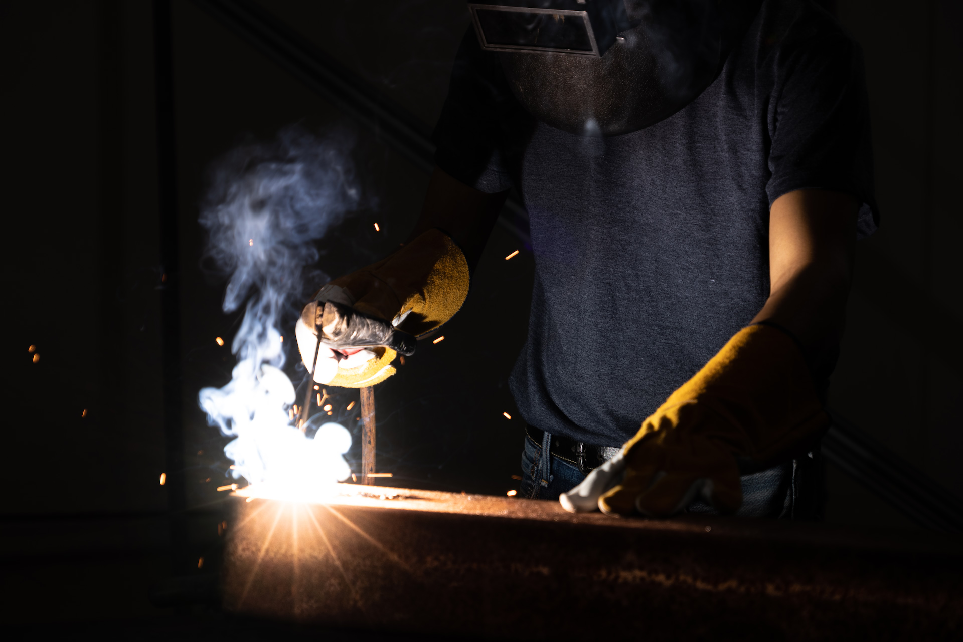 Hands in heavy gloves welding at a workbench in the dark, smoke rising