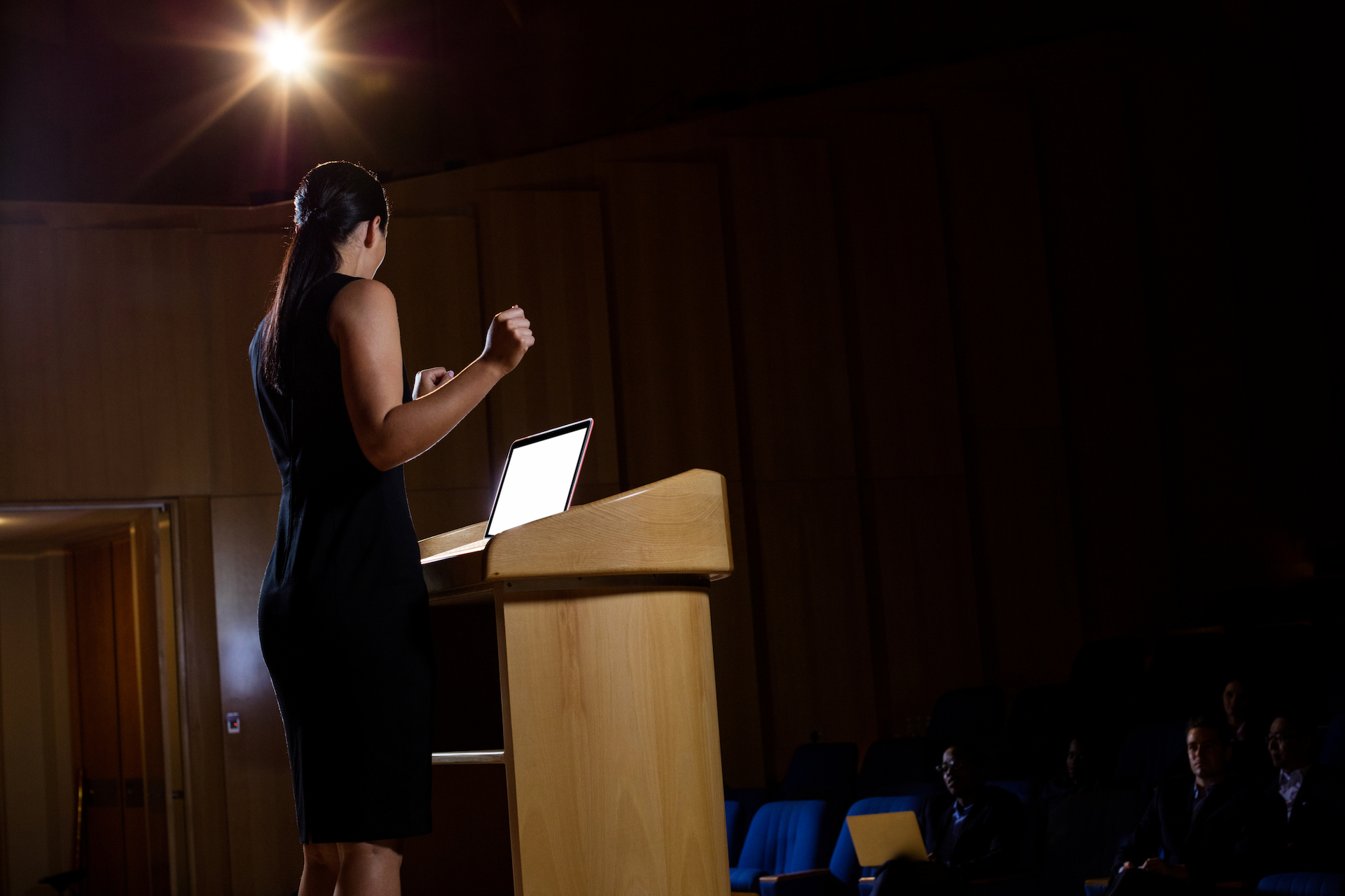 A speaker at a lectern with a laptop, addressing a darkened auditorium