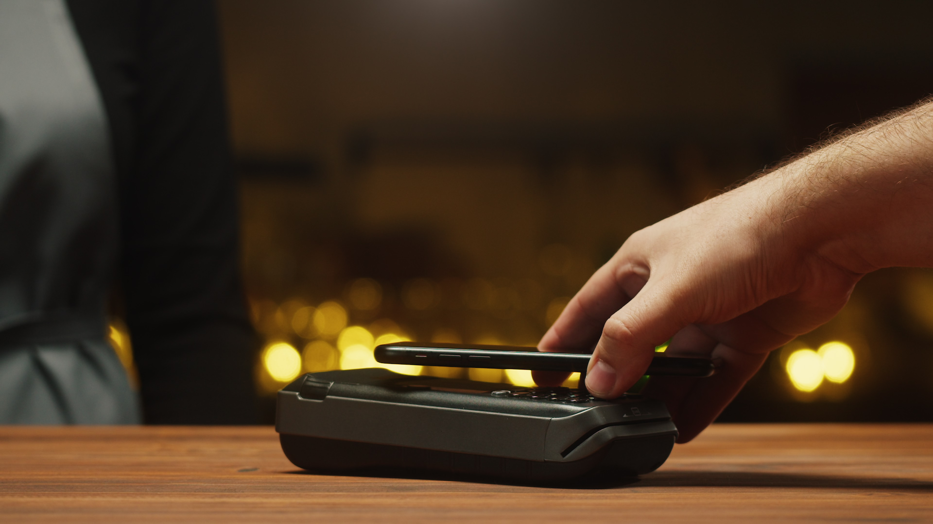 A hand holding a phone to a card terminal to pay, warm lights behind